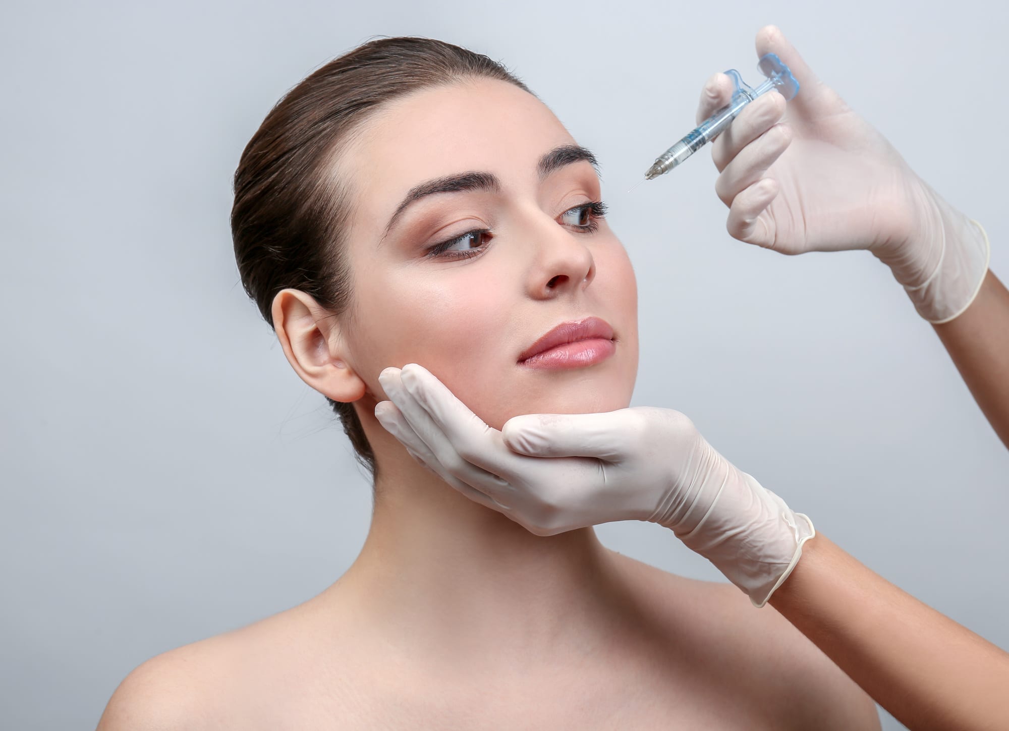 Beautiful young woman receiving filler injection in face, on light background